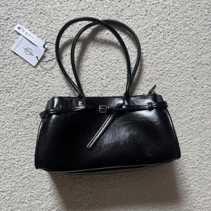 Svart handväska från Stradivarius - Stilren svart handväska från Stradivarius i glansigt syntetmaterial. Väskan har dubbla handtag, dekorativt bälte framtill och en klassisk, rektangulär form. Perfekt för dig som gillar minimalistisk och trendig design.