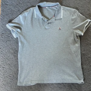 Grå pikétröja från Moncler - Snygg grå pikétröja från Moncler med klassisk krage och vita ränder på ärmslut och krage. Tröjan har korta ärmar och Moncler-logga på bröstet. Tillverkad i mjuk bomull som känns skön mot huden. Perfekt för en clean och stilren look.