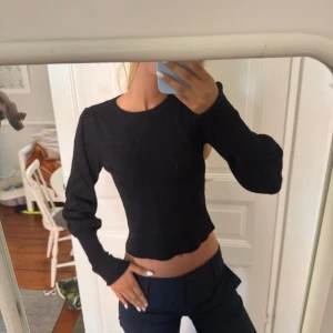 Svart stickad långärmad tröja - En svart stickad tröja med lång ärm och rund hals. Modellen är figurnära med lätt croppad passform och diskret vågig nederkant. Perfekt för dig som gillar en enkel men stilren look.