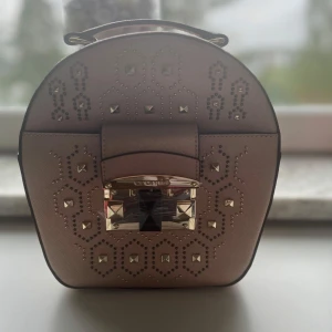 Beige axelväska från Cromia med nitar - Rundad beige axelväska från Cromia med coola guldiga nitar och dekorativa mönster framtill. Väskan har ett snyggt spänne i guld och svart, samt dragkedja upptill. Tillhörande axelrem medföljer. Perfekt för dig som gillar detaljer och unik design.