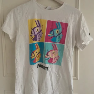 KÖP FRÅN MIN VINTED - Vit t-shirt från Fortnite med ett stort, färgglatt tryck av fyra llamor i pop art-stil på bröstet. T-shirten har korta ärmar och Fortnite-logga tryckt på ena ärmen. Tillverkad i mjuk bomull, perfekt för gaming eller chill.