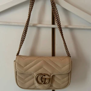 Beige quiltad axelväska  - Säljer en beige axelväska med klassiskt GG-spänne i guld. Väskan är quiltad i skinn och har en snygg kedjerem i metall. Perfekt storlek för det viktigaste och ger en lyxig touch till din outfit.