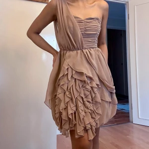 Champagnefärgad volangklänning H&M - Supersnygg kort klänning från H&M i champagnefärgad ton. One shoulder-modell med draperad överdel och massor av volanger på kjolen. Tillverkad i ett lätt, flowy material som ger en lyxig känsla. Perfekt för dig som vill sticka ut med en unik look.