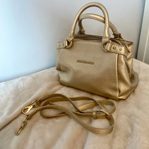 Guldig handväska från Roccobarocco - Snygg guldig handväska från Roccobarocco i präglat skinn. Inga defekter finns, aldrig använd av mig tyvärr 🥲💗. Väskan har både handtag och avtagbar axelrem, dragkedja upptill och coola metalldetaljer. 
