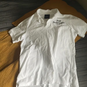 Vit Peak Performance pikétröja - Klassisk vit pikétröja från Peak Performance med svart broderad logga på bröstet. Tröjan har korta ärmar, krage och knappar framtill. Perfekt för en clean och sportig stil. Tillverkad i mjuk bomull som andas.