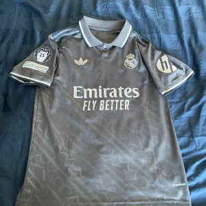 Real Madrid Mbappé grå fotbollströja - Snygg grå Real Madrid fotbollströja från Adidas med Mbappé och nummer 9 på ryggen. Tröjan har korta ärmar, krage och diskret mönster i tyget. Broderade klubb- och sponsorloggor samt Champions League-märke på ärmen. Tillverkad i lätt och ventilerande material.