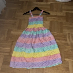 Pastellrandig långklänning med smock - Superfin långklänning i pastellfärger med breda ränder i lila, rosa, gul, grön och blå. Klänningen har smock upptill och smala axelband. Perfekt för sommarens varma dagar och ger en färgglad och lekfull vibe.