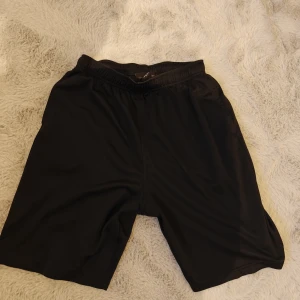 Svarta träningsshorts från Bauer - Svarta träningsshorts från Bauer med elastisk midja och enkel, loose passform. Tillverkade i polyester som andas, perfekta för sport eller gym. Klassisk look utan mönster eller extra detaljer, bara riktigt sköna shorts för dig som gillar att röra på dig.