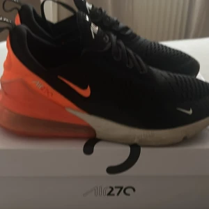 Nike Air Max 270 svart/orange strl 44 - Nike Air Max 270 sneakers i svart mesh med orange detaljer och stor synlig air-enhet i hälen. Snygg design med vita och orangea loggor, snörning och platt sula. Perfekta för dig som gillar streetstyle och vill sticka ut med färg. Är som nya använt 5 gånger köpte för 1 månad sedan 