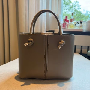 Beige handväska Zara - Stilren beige handväska i skinn med två handtag och snygga knutdetaljer framtill. Väskan har magnetlås och ett innerfack med dragkedja för extra säkerhet. Perfekt storlek. Ett längre band tillkommer så man även kan ha den över axeln. Aldrig använd, nyskick.