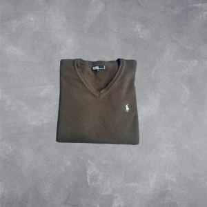 Brun v-ringad tröja Polo Ralph Lauren - Snygg brun v-ringad tröja från Polo Ralph Lauren med klassisk broderad logga på bröstet. Tröjan har långa ärmar, ribbade muddar och är tillverkad i mjuk bomull. Perfekt för lager-på-lager och enkel att matcha med jeans eller chinos.
