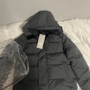Canada Goose black label - Säljer en grå dunjacka från Canada Goose med huva och dragkedja. Jackan har en broderad logotyp på ärmen och är perfekt för kalla dagar. Den är i storlek S och har en klassisk pufferdesign. Bra skick  perfekt till vinter och kalla dagar tag jack påsen med följer skriv om ni undrar något kan gå ner i pris såklart vid köp 