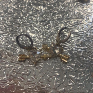 Guldiga örhängen med hjärtan och pil - Snygga örhängen i guldton med hjärtformade hängen och pil-detaljer. Framsidan har glittrande stenar i hjärtform, medan baksidan visar ett utskuret hjärta i metallen. Perfekta för dig som gillar unika accessoarer med romantisk vibe.