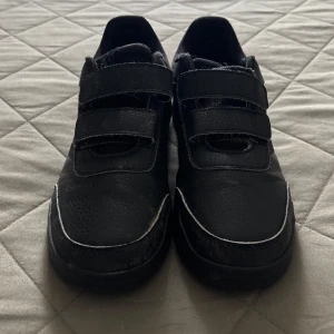 Svarta sneakers från Adidas med kardborre - Svartas sneakers från Adidas med två breda kardborreband för enkel påtagning. Skorna har diskreta ränder på sidan, rund tå och platt sula.  Skriv för fler frågor eller bilder i chatten tack!