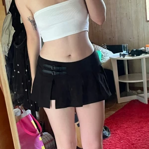 Svart plisserad minikjol med spännen - Supersnygg svart kortkjol med plisserad design och två coola spännen i midjan. Kjolen har inbyggda shorts under för extra komfort och passar perfekt till en edgy eller sportig stil. Materialet känns mjukt och följsamt.