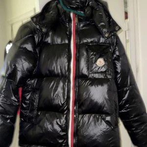 Svart Moncler pufferjacka M - Säljer en svart, glansig pufferjacka från Moncler med avtagbar huva och klassiska Moncler-detaljer. Jackan har dragkedja med dubbla zippar, ficka med logga på bröstet och röd-vit-blå rand längs dragkedjan. Perfekt för kalla dagar och riktigt snygg streetstil.