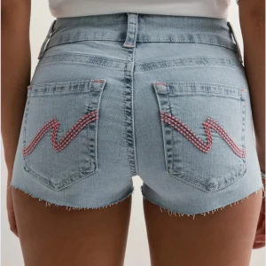 Ljusblå jeansshorts med rosa brodyr - Snygga ljusblå jeansshorts med rå kant och cool rosa brodyr på bakfickorna. Shortsen har låg midja och klassisk femficksdesign. Perfekta för varma dagar när du vill ha en avslappnad men trendig look.