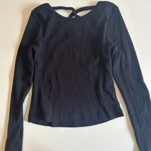 Svart ribbad topp med twistad rygg KappAhl - Svart långärmad topp från KappAhl med ribbad struktur och snygg twistad detalj i ryggen. Modellen är figurnära och har en enkel, stilren look som passar till mycket. Perfekt barn som vill vara lite fina men samtidigt vardagliga. 