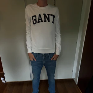 Vit sweatshirt från GANT - Säljer en stilren vit sweatshirt från GANT med svart broderad logga över bröstet. Tröjan har rund halsringning och långa ärmar, perfekt för en clean och klassisk look. Passar dig som gillar enkel och snygg streetstyle.