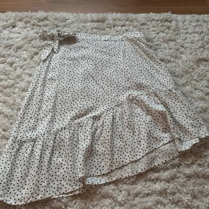Vit prickig kjol med volang - Vit prickig sneskuren kjol med en knyt rosett på ena midjan. Väldigt fin att matcha med exempelvis en svart eller vit tröja! Den ger en basic outfit en liten extra touch. Används ett par gånger men inga tecken på slitage! 