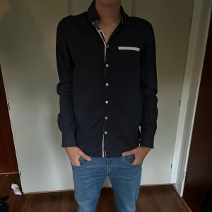 Mörkblå skjorta från Savvy Citizen - Snygg mörkblå skjorta från Savvy Citizen med vita detaljer vid knappslå och bröstficka. Skjortan har långa ärmar och klassisk krage, perfekt för en stilren look. Tillverkad i mjuk bomull som känns skön mot huden.