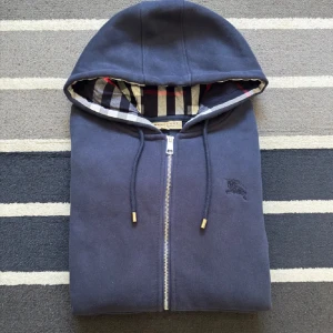 Marinblå hoodie från Burberry - Snygg marinblå hoodie från Burberry med dragkedja och klassiskt rutigt foder i huvan. Tröjan har snörning i huvan, broderad logga på bröstet och guldfärgade detaljer på snörena. Perfekt för dig som gillar stilrena och exklusiva plagg.