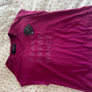 Mörkrosa Peak Performance t-shirt XS - Snygg mörkrosa t-shirt från Peak Performance i bomull med tryckt logga vertikalt framtill. Modellen är ärmlös med rund halsringning och har en clean, sportig vibe. Perfekt för dig som gillar färg och vill sticka ut lite extra. Står XS, passar även S