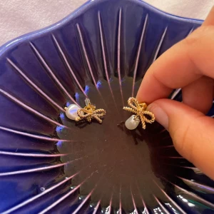 Guldiga örhängen med rosett och pärla - Supersöta örhängen i guld med tvinnad rosett och hängande vit pärla. Perfekta för att ge din look en gullig och elegant touch. Rosetten är detaljerad och pärlan har en naturlig, oregelbunden form som gör dem unika. Helt nya aldrig använd 