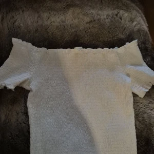 Vit offshoulder topp från H&M - Supersöt vit offshoulder topp från H&M med smockad struktur och volangkant upptill. Toppen är ärmlös och har en tight passform som framhäver axlarna. Perfekt för varma dagar när du vill ha en fräsch och stilren look.