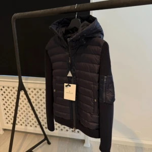 Mörkblå Moncler pufferjacka med huva - Snygg mörkblå pufferjacka från Moncler med stickade ärmar och vadderad front. Jackan har en stor huva, dragkedja framtill och coola detaljer som ficka med dragkedja på ärmen samt Moncler-logga. Perfekt för dig som vill ha en stilren och sportig look. 10/10 skick!