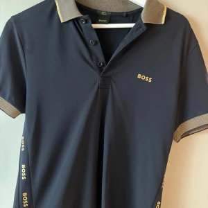 Svart pikétröja från BOSS med gulddetaljer - Svart pikétröja från BOSS med guldfärgade detaljer och logga på bröstet och ärmarna. Tröjan har klassisk krage i kontrasterande beige/guld, korta ärmar och knäppning framtill. Slim fit-modell i stretchigt material som sitter snyggt på kroppen. STRL S-Passar för de som pendlar mellan S & M i strl! Använd endast en gång!👌🏼