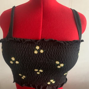 Svart smockad linnetopp med broderade blommor - Supersöt svart smockad linnetopp med tunna axelband och små broderade vita och gula blommor. Toppen har volangkant upptill och nedtill för en extra gullig vibe. Perfekt för att lyfta vilken outfit som helst med en touch av sommarfeeling.