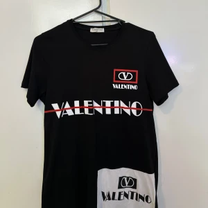 Svart Valentino t-shirt med tryck - Cool svart t-shirt från Valentino med stort vitt logotryck och röd linje över bröstet. Extra detalj med vit patch och logga nedtill. Klassisk rund hals och korta ärmar. Perfekt för dig som gillar streetstyle och statement-plagg.