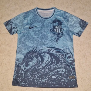 Blå FC Barcelona x Nike draktröja - Unik blå FC Barcelona fotbollströja från Nike med detaljerat drakmönster över hela plagget. Tröjan har rund hals, korta ärmar och klubbens emblem på bröstet. Tillverkad i lätt och ventilerande polyester, perfekt för dig som vill sticka ut på planen.