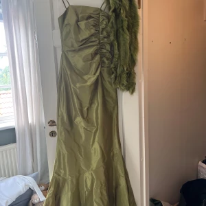 Olivgrön långklänning från Angie samt matchande fjädersjal  - Snygg olivgrön långklänning från Angie med smala axelband och figurnära passform. Klänningen har en glansig yta, rynkade detaljer framtill och dragkedja i ryggen. Perfekt för dig som vill sticka ut med en elegant och unik look.
