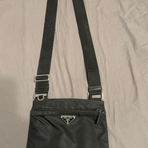 Prada Nylon Crossbody - Prada Nylon Axelväska - Äkta Prada axelväska i klassisk svart nylon med ikoniska triangelloggan framtill. Enkel, stilren och perfekt för både vardag och resa. Justerbart axelband. Modellen är unisex och väldigt lätt att bära.  Väskan är i fint begagnat skick (7–8/10) – inga stora defekter, bara normalt slitage.  En tidlös favorit som inte tillverkas längre!