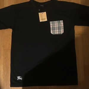 Svart Burberry t-shirt med rutig ficka - Svart t-shirt från Burberry med klassisk rutig ficka på bröstet och broderad vit logga nertill. T-shirten har korta ärmar och är gjord i mjuk bomull. Snygg och stilren med en touch av Burberrys ikoniska design.