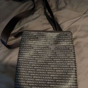 Armani Exchange axelväska med logomönster - Snygg svart axelväska från Armani Exchange med justerbart band och dragkedja upptill. Väskan har ett stilrent heltryck med loggan 'Armani Exchange' över hela ytan. Perfekt storlek för det viktigaste och en clean look till vardagsoutfiten.