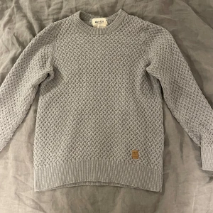 Grå stickad tröja från NN07 - Snygg grå stickad tröja från NN07 med coolt vävt mönster och ribbade muddar. Tröjan är i 100% bomull och har en klassisk rund halsringning. Perfekt för lager-på-lager och chill stil.