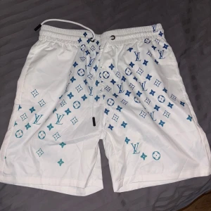 Louis Vuitton vita badshorts med monogram - Snygga vita badshorts från Louis Vuitton med blått och turkost monogrammönster. Shortsen har elastisk midja med snörning och klassisk LV-logga tryckt över hela tyget. Perfekta för stranden eller poolen, riktigt lyxig vibe och skön passform. Storlek: L men passar M också.