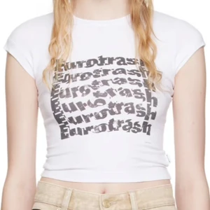 Eytys Eurotrash Baby Tee - Säljer en vit croppad t-shirt med svart texttryck 'Eurotrash' i flera lager på bröstet. T-shirten har korta ärmar och en tight passform som framhäver midjan. Perfekt för dig som gillar edgy och trendiga plagg.