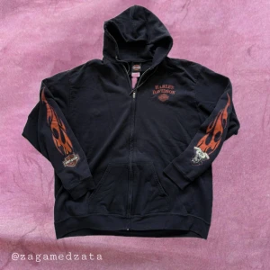Vintage Harley-Davidson hoodie med flames - Svart hoodie från Harley-Davidson med dragkedja och huva. Snygga orange flames på båda ärmarna och stort tryck på ryggen med logga och text. Perfekt för dig som gillar bikerstil och vill sticka ut. Mjuk bomullskänsla och coola detaljer. Storleken har nöts bort på lappen men upskattar den till en storlek XL.