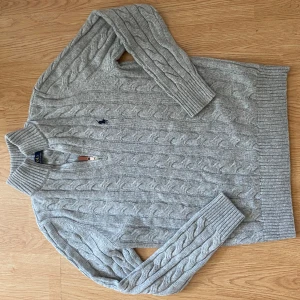 Grå kabelstickad polotröja Gant - Snygg grå kabelstickad polotröja från Ralph raulen med halv dragkedja och broderad logga på bröstet. Tröjan har ribbade muddar och hög krage, perfekt för kyliga dagar. Klassisk och stilren design som funkar till det mesta.