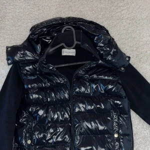 Moncler mörkblå pufferjacka med huva - Snygg mörkblå pufferjacka från Moncler med glansig yta och avtagbar huva. Jackan har quiltade partier framtill och på ryggen, samt stickade ärmar och sidor för extra komfort. Dragkedja framtill och praktiska fickor med tryckknappar.