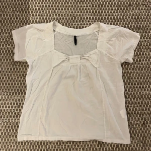 Vit blus med rosett från Naf Naf - Supersöt vit blus från märket Naf Naf med korta ärmar och en stor rosett framtill. Blusen har en fyrkantig ringning och är gjord i ett tunt, luftigt material som känns perfekt för varmare dagar. Den fladdrar fint och vintage från 2000-talet men tyvärr finns ett litet hål på höger axel. Skriv gärna om ni har några frågor!!