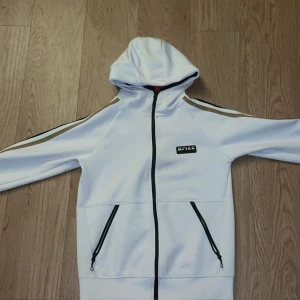 Vit hoodiejacka från Hugo boss med dragkedja - Vit hoodiejacka från Hugo boss med svarta och bruna ränder längs ärmarna. Jackan har hel dragkedja framtill, två fickor med dragkedja och en huva. Sportig look med stilrena detaljer och logga på bröstet. Perfekt för dig som gillar streetwear.