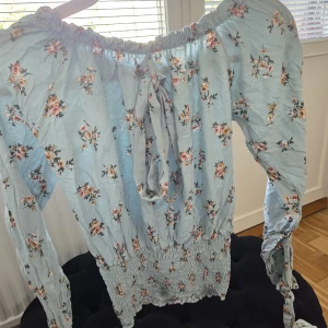 Blommig offshoulder blus i ljusblått - Supersöt ljusblå blus med blommigt mönster och offshoulder-modell. Smockad midja och ärmslut ger en snygg passform. Långa ärmar och knytdetalj framtill. Perfekt för vår och sommar när du vill känna dig extra fin.