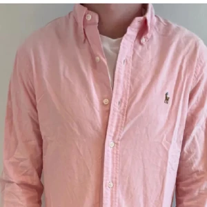 Rosa skjorta från Ralph Lauren - Säljer en klassisk rosa skjorta från Ralph Lauren med button-down krage och broderad logga på bröstet. Skjortan har långa ärmar och knäppning framtill. Perfekt för dig som gillar stilrena och tidlösa plagg.