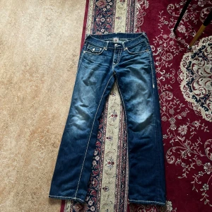 True Religion blå jeans med kontrastsömmar - Säljer ett par klassiska blå jeans från True Religion med tydliga vita kontrastsömmar och ikoniska bakfickor med lock och knapp. Jeansen har en rak passform och är tillverkade i kraftigt denim. Märkeslapp bak och snygg tvättad look.