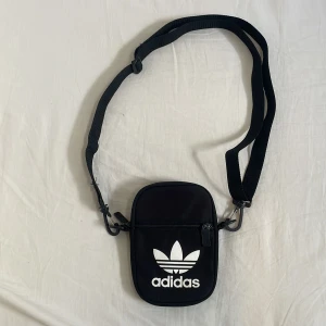 Svart axelväska från Adidas - Snygg svart axelväska från Adidas med klassisk vit logga framtill. Väskan har justerbar rem, dragkedja och flera fack inuti för enkel organisering. Perfekt storlek för mobil, plånbok och andra småsaker. Tillverkad i slitstark polyester.